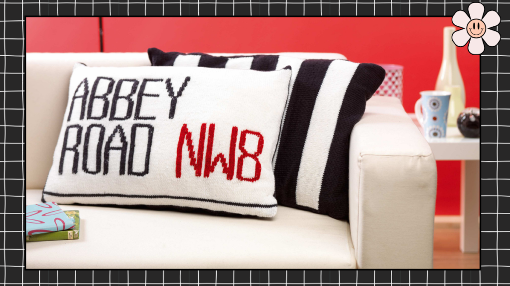 Two knitted cushion covers on a white sofa — Abbey Road NW8 street sign cushion in black, white and red, alongside a black and white striped zebra crossing cushion, knitting pattern by Jane Burns